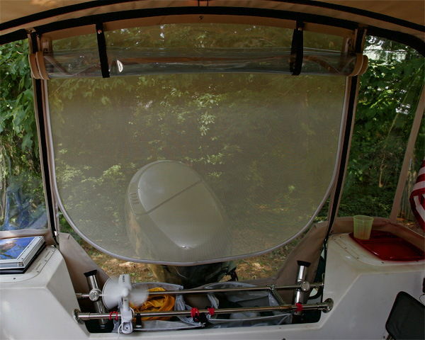 Looking aft with camperback window rolled up to expose screening. We are told that the screening blocks visibility from the rear, although we see fine from inside. Fan clamps to transom bars and makes a big difference in hot weather. Nylon mesh bags sit down inside bars to hold drinks, water jugs, and propane cylinders kept in a porous nylon bag. Any leakage should go out of the splash well outlet. We normally lash a large cooler on the bars when underway to free up floor space. Rear panel goes behind lazerettes to free up significant space.