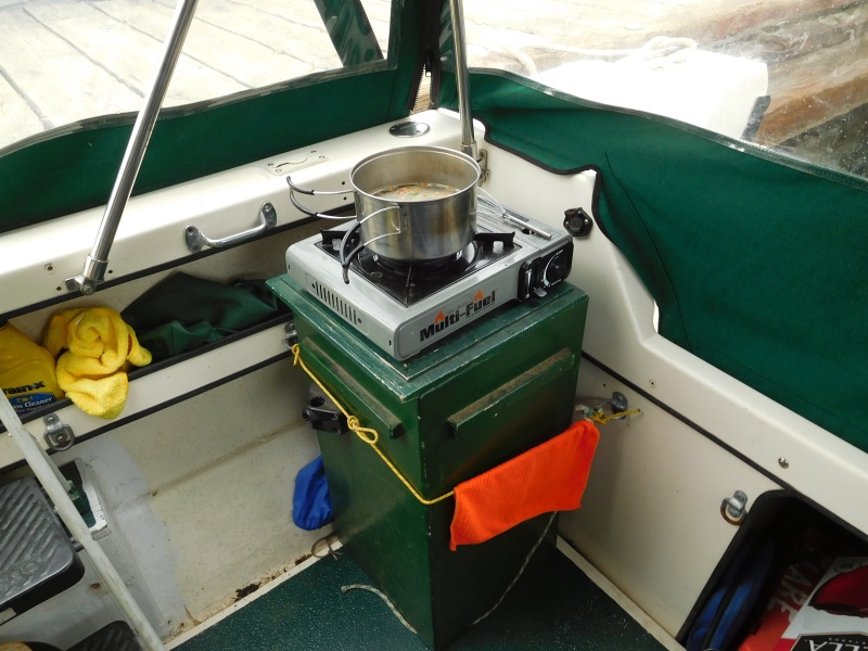 Outside Cooking.
Built box to hold stove on top for cooking and inside for travel.  Also holds fuel & many other bits of gear.  Rope holds  in place when trailering.  Note "D" rings mounted around inside  of cockpit to tie in gear.  Black 6 sided thing near front corner of stove is cap over 12 volt electric inlet/ outlet for line from tow vehicle  to power house battery when trailering or to provide 12 volt power in cockpit.