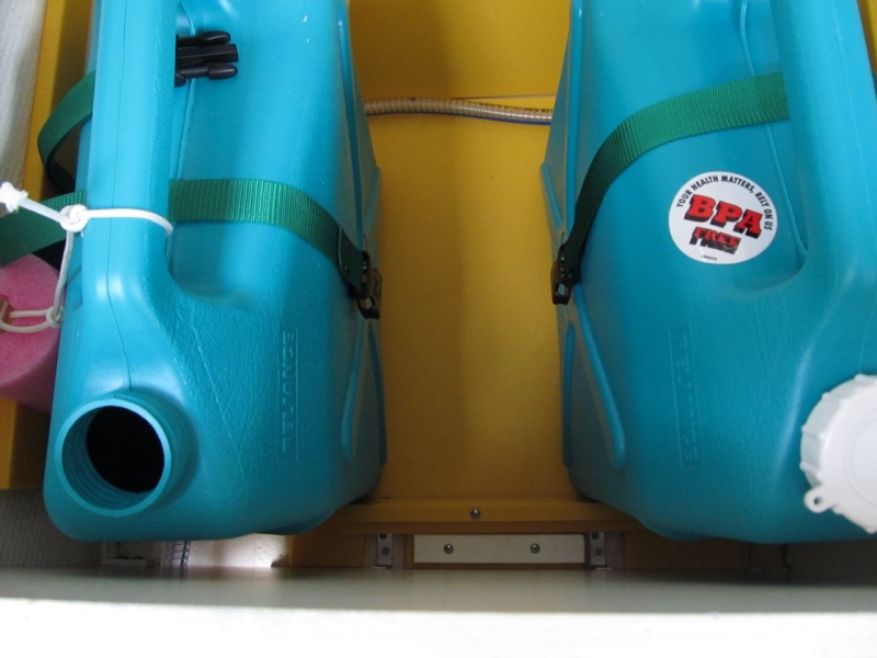 The yellow Starboard? platform with white cleats was set up for my original water tank, which then sprung a leak.  I'm planning to get a new/permanent tank, but in the meantime set up this system.  A temporary full width Starboard cleat holds the jugs off the upper/inboard locker side.  Three jugs just fit, with storage outboard (behind them).
